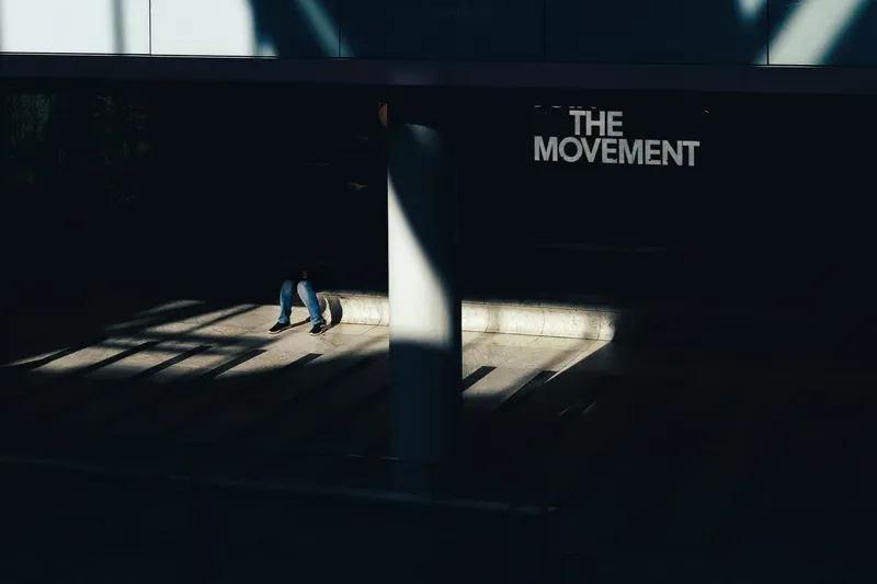 A seated figure's legs are lit by a stray beam of light in an otherwise dark hall beneath a sign reading 'THE MOVEMENT'.