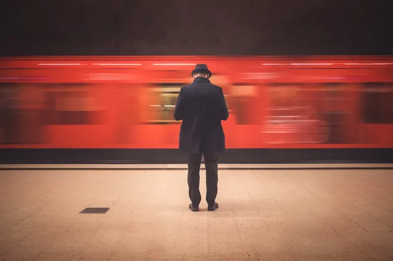 A train blurs past in red light as a man in a hat stands still on the platform.