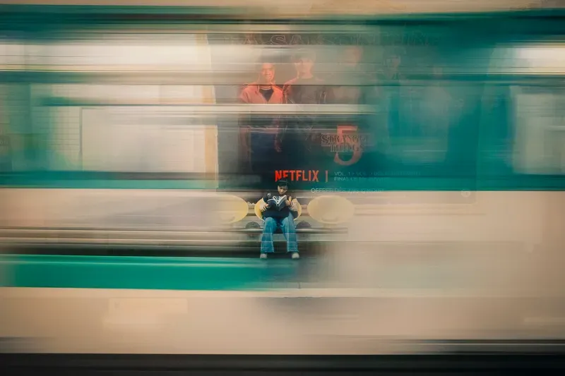 A person sits reading a book on a station bench, motion blur streaking the arriving train and a Stranger Things ad on the wall behind them.