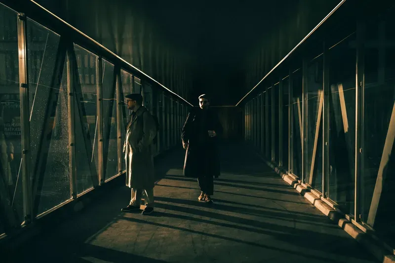 Two men in trench coats and sunglasses stand in a dark corridor cut by a dramatic diagonal beam of golden light.