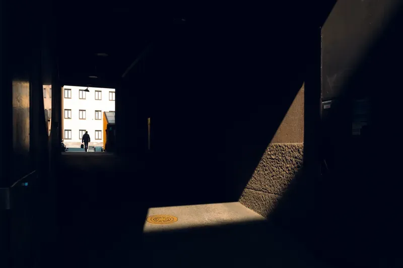 A dark concrete underpass with a bright doorway at the far end, a small figure silhouetted in it, and a shaft of sunlight falling across a manhole cover.