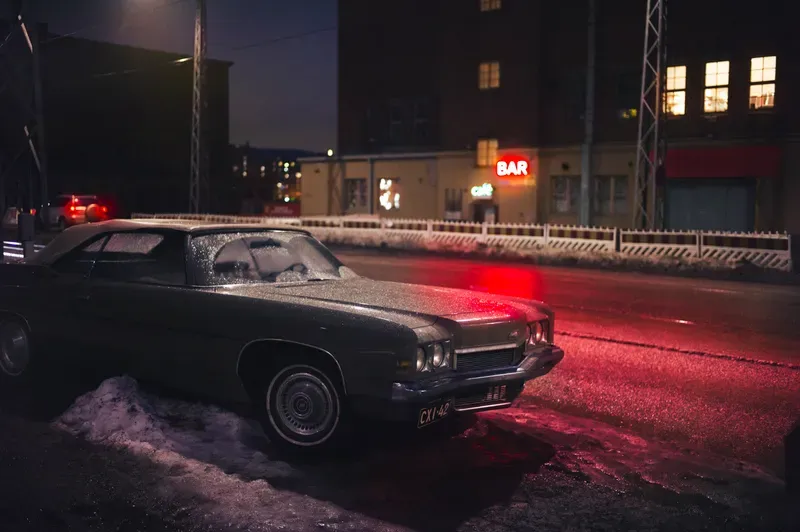 A frost-covered vintage car sits parked on a snowy street at night, a glowing red 'BAR' neon sign lighting the building beyond.