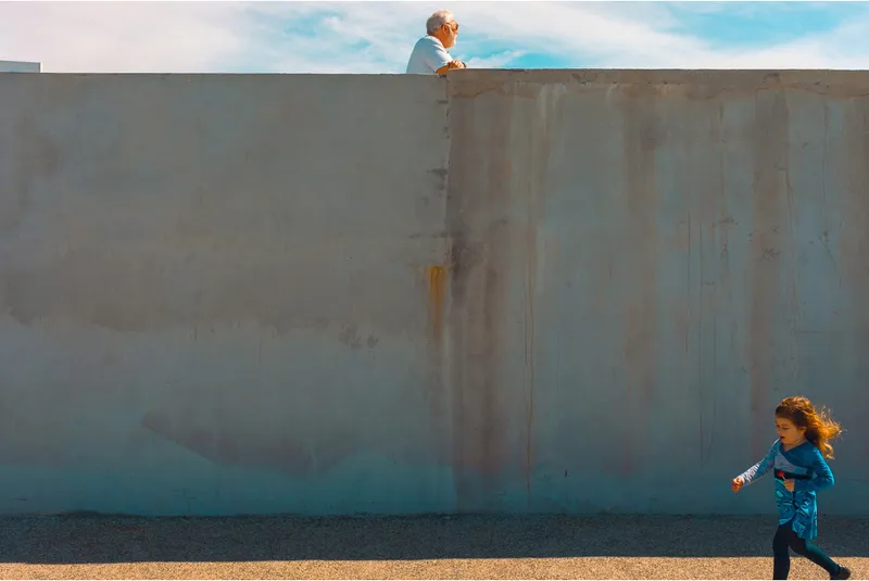 An old man peers over a rusted concrete wall as a young girl runs past below in the sunshine.