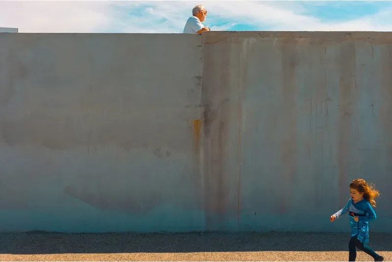 An old man peers over a rusted concrete wall as a young girl runs past below in the sunshine.