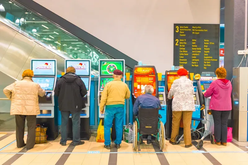 A row of shoppers seen from behind play betting machines beside an escalator, one of them in a wheelchair, a shopping-centre directory board on the wall above.