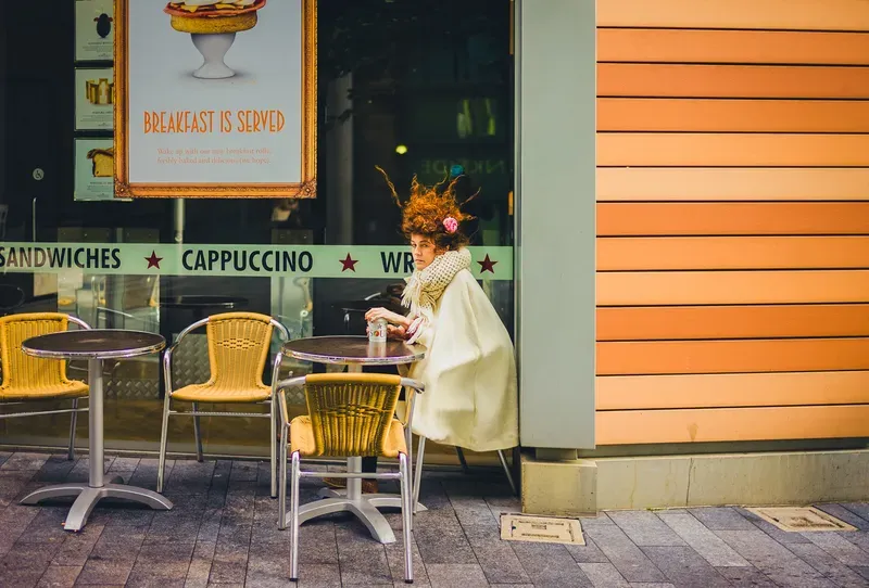 A woman with wind-tossed curly hair and a cream cape sits at a cafe table, glancing back over her shoulder beside a breakfast poster.