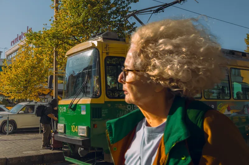 A woman with windswept grey-blonde curls and tortoiseshell glasses looks off to one side in strong autumn sun, a green and yellow Helsinki tram and yellowing trees behind her.