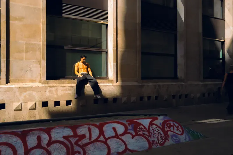 A shirtless man sits on a stone window ledge above a graffitied wall, looking down at the empty street below.