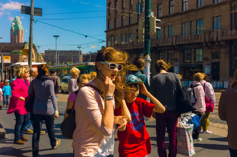 A girl in sunglasses with windswept hair walks through a crowded city square, a tram and station tower visible behind her.