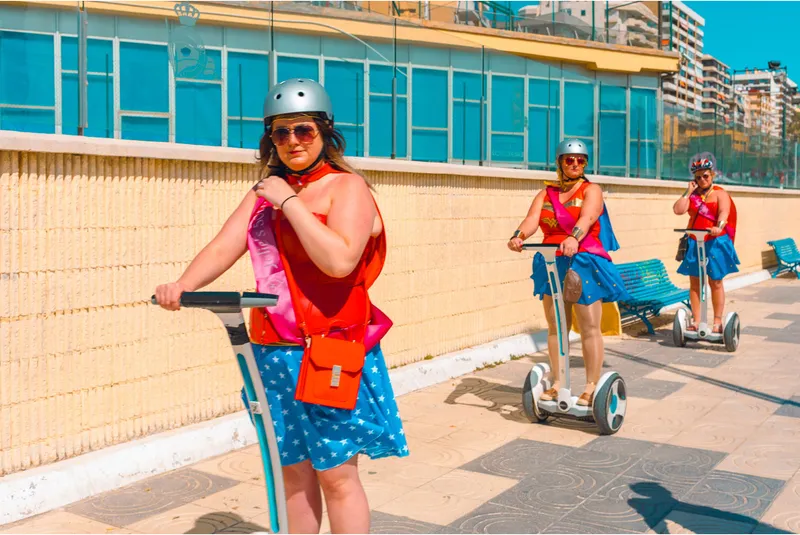 Women dressed as Wonder Woman ride Segways along a seaside promenade in matching costumes and helmets.