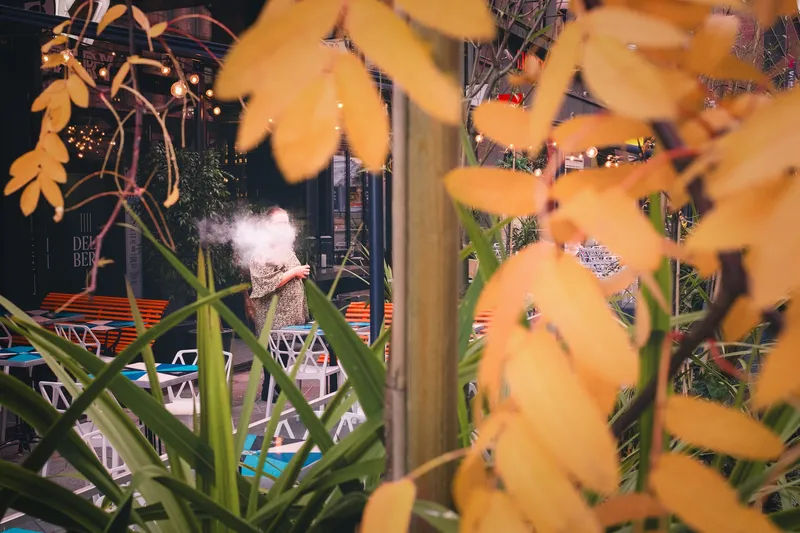 Golden autumn leaves frame a woman exhaling vapor outside a café, string lights glowing behind her.