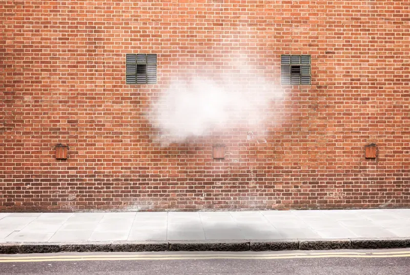 A cloud of steam drifts up a red brick wall between two small shuttered windows above an empty sidewalk.