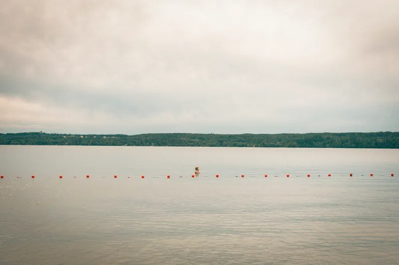 A lone swimmer's head is just visible in a still, pale lake marked by a line of buoys.