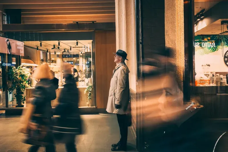 A woman in a trenchcoat is caught mid-stride in motion blur past a brightly lit shopfront window at dusk.