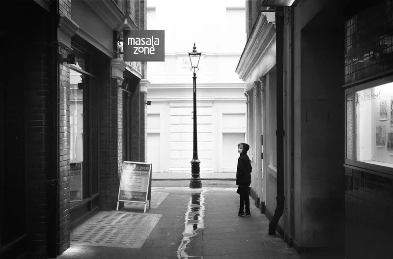 A child in a hooded coat stands alone in a narrow London alley beside an Indian restaurant sign and a lit street lamp.