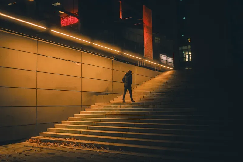A man descends a warmly lit staircase past a glowing red neon sign in a Helsinki underpass at night.
