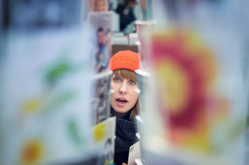 A woman in an orange beanie looks through a rack of postcards, her face framed between blurred colorful cards.