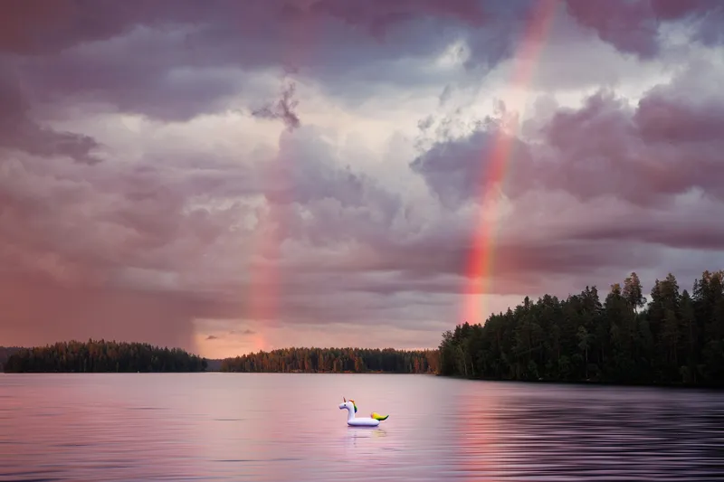 A double rainbow arcs over a calm lake at dusk, an inflatable swan floating alone near the shore.