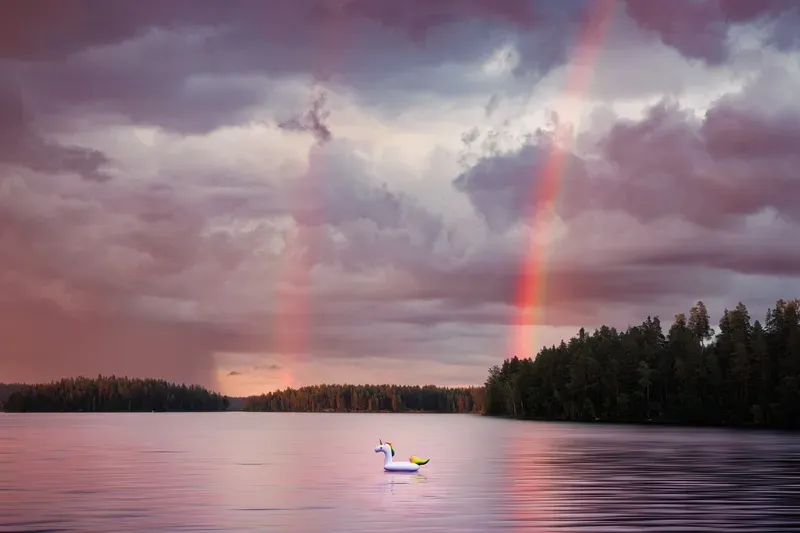 A double rainbow arcs over a calm lake at dusk, an inflatable swan floating alone near the shore.