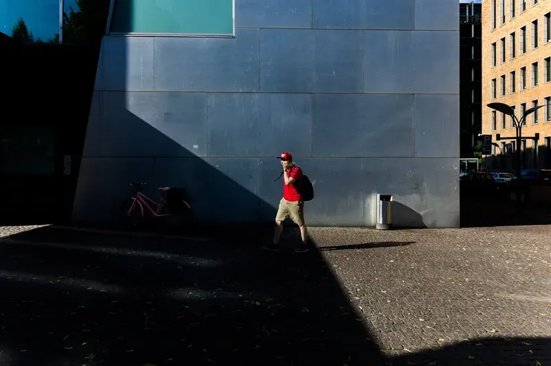 A man in a red cap walks across a sunlit plaza, a long shadow cast beside a curved concrete building.