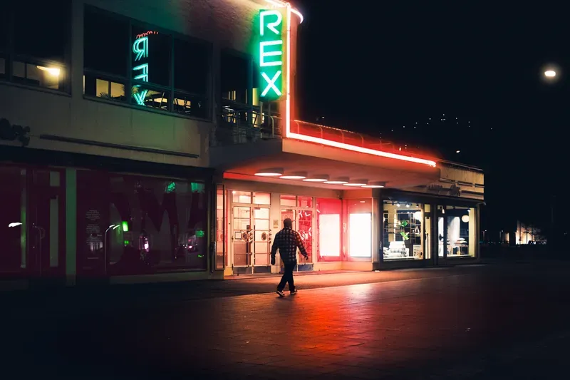 A man walks past a cinema's green and red neon signage reflected on the wet pavement of an empty square at night.