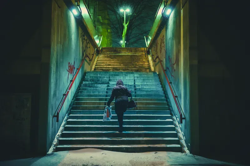 A figure climbs a snow-dusted staircase at night, lit by green and amber lights beneath bare winter trees.