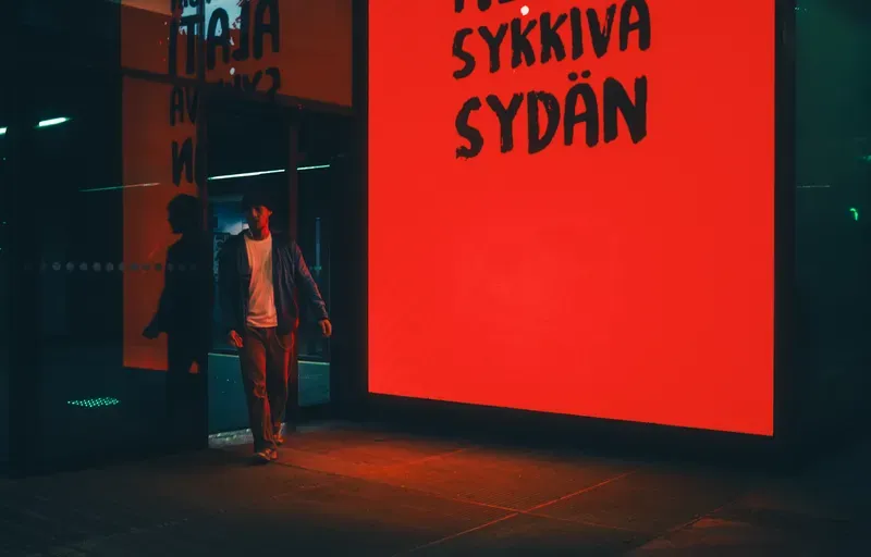 A man walks out of a doorway lit red by a glowing sign reading Sykkivä Sydän beside a shop window at night.