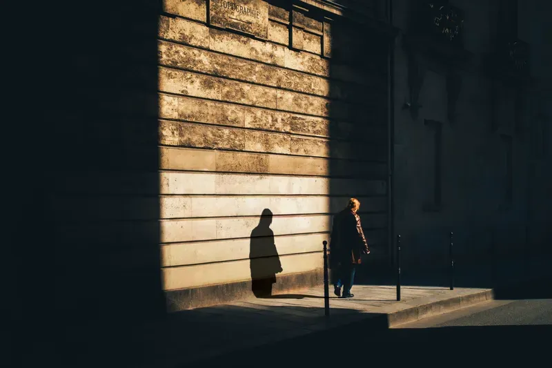 Two figures stand in a sharp diagonal of light and shadow cast across a ridged building facade.