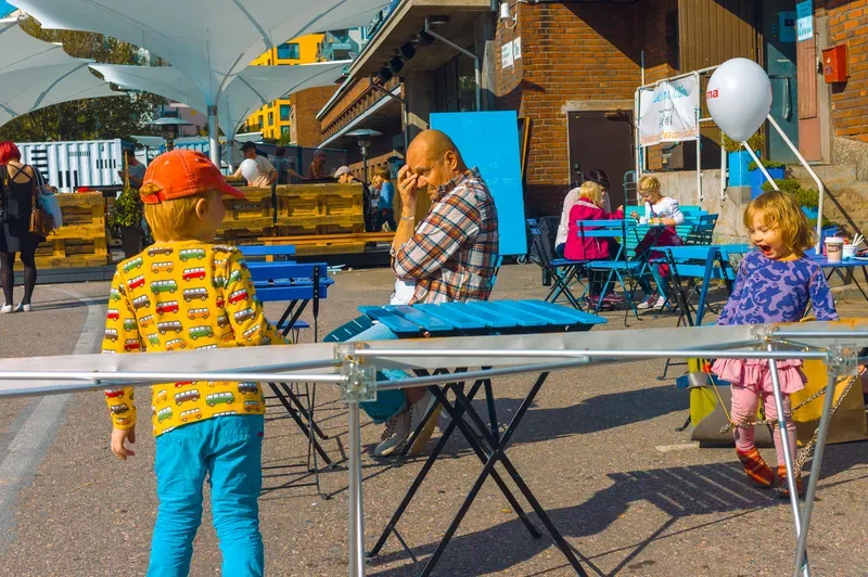 A boy in an orange cap and a yellow top printed with little buses stands in a sunlit street café area, a man with his hand to his face at a blue table and a small girl in purple laughing at the right edge.