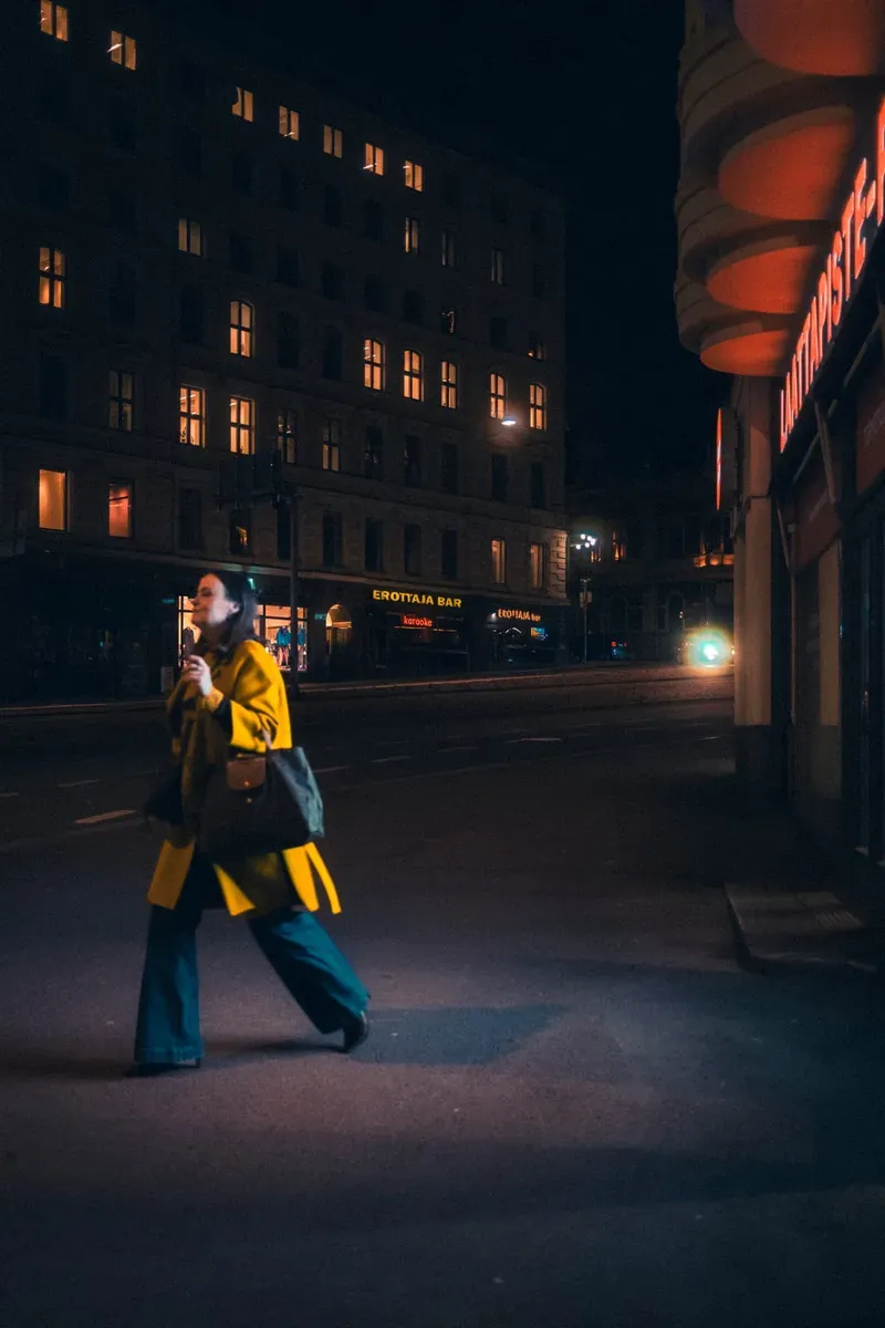 A woman in a yellow coat and teal trousers strides across a dark street at night, lit windows glowing in the building behind her.