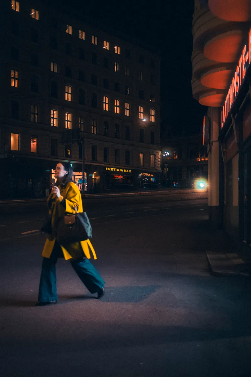 A woman in a yellow coat and teal trousers strides across a dark street at night, lit windows glowing in the building behind her.