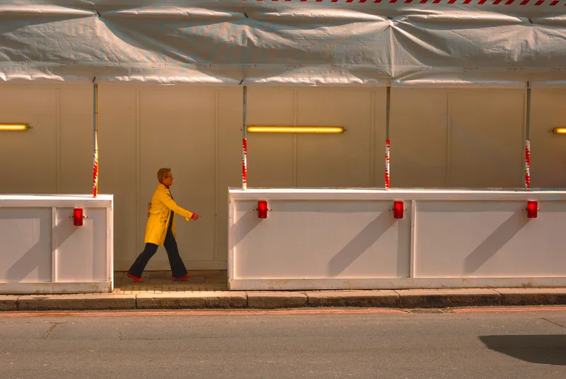 A woman in a yellow coat and red shoes walks past a row of red hazard lights on a construction hoarding.