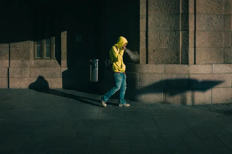 A figure in a yellow jacket walks past a wall, casting a long shadow across the sunlit pavement.